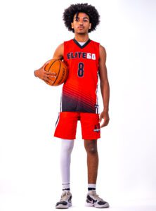 A boy with curly hair wearing a red basketball jersey and holding a ball near his armpits