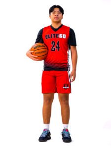 An Asian boy in a red basketball jersey holding a ball on his side