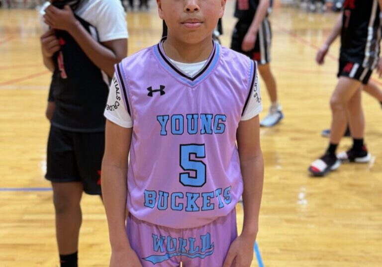 Young basketball player wearing a pink and blue jersey stands on court.